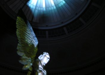 A sculpture of a winged figure under a skylight dome with dramatic lighting illuminating its form