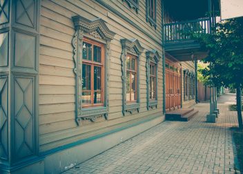 Vintage wooden building facade with ornate window frames and a shaded pavement walkway in a sunny setting