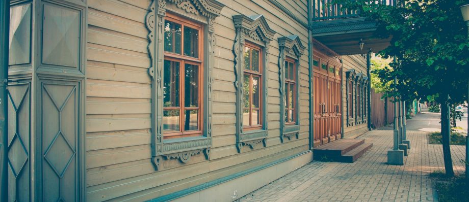 Vintage wooden building facade with ornate window frames and a shaded pavement walkway in a sunny setting
