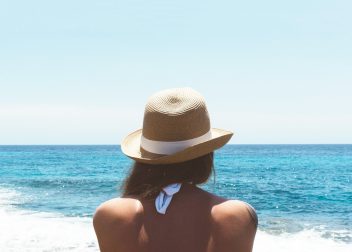Woman in sun hat gazing at the ocean, enjoying a sunny beach day, with blue sky and sea horizon in the background
