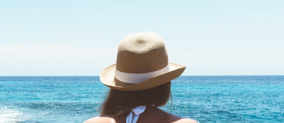 Woman in sun hat gazing at the ocean, enjoying a sunny beach day, with blue sky and sea horizon in the background