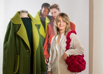Fashion designer posing with green coat on mannequin and holding bold red bag at fashion exhibit display