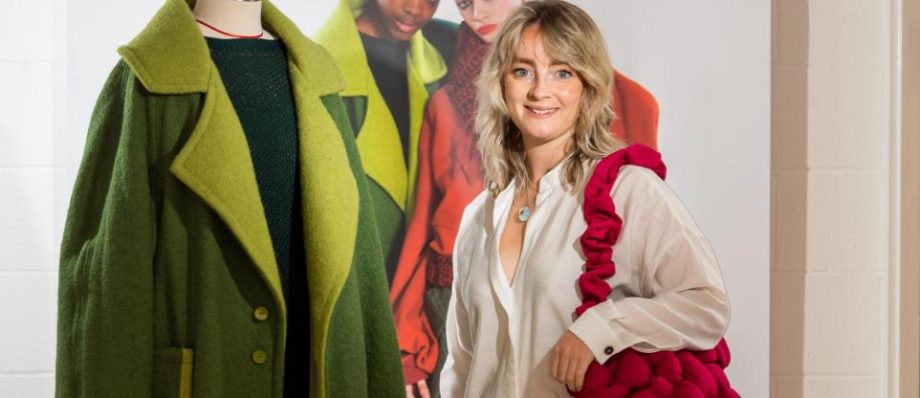 Fashion designer posing with green coat on mannequin and holding bold red bag at fashion exhibit display
