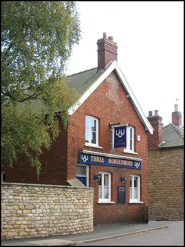 Historic UK pub with brick façade named Three Horseshoes, featuring blue signage and surrounded by rustic buildings