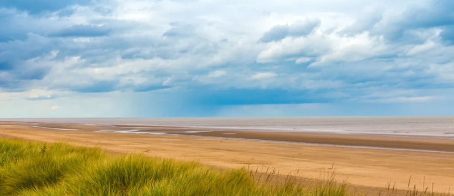Expansive sandy beach with grassy dunes and a dramatic cloudy sky, peaceful coastal scene in the UK