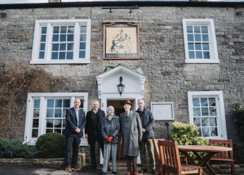 Group of people standing outside Assheton Arms pub in front of a stone building with windows and door