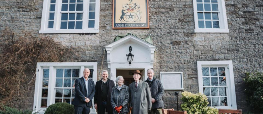 Group of people standing outside Assheton Arms pub in front of a stone building with windows and door