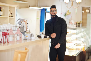 Man in a black jacket standing by a cafe counter, surrounded by cosy decor and soft lighting