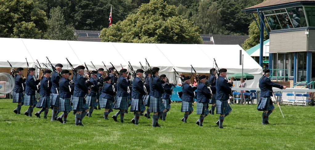 Scottish pipe band in traditional kilts performing at an outdoor event with lush greenery in the background