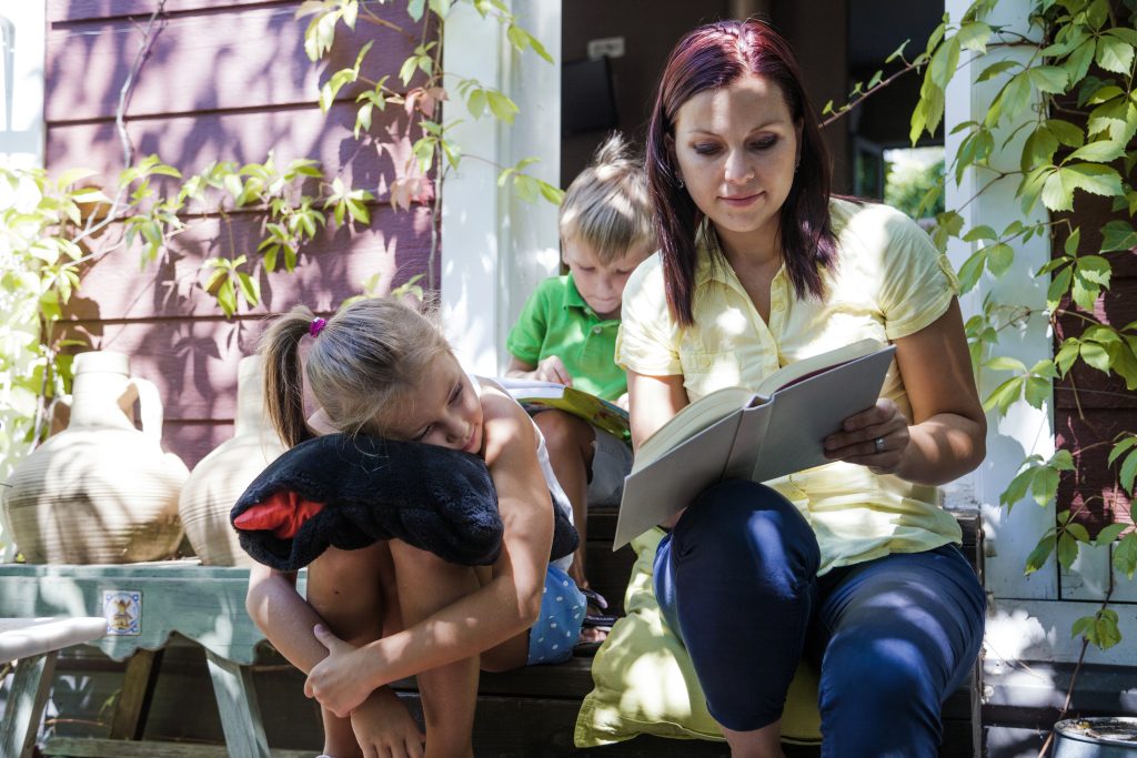 Mother reading book on porch with two children, girl hugging plush toy, summer day scene, family bonding outdoors
