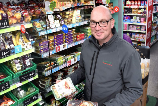 Man shopping for groceries in a store aisle, holding ready meals amidst fresh produce and packaged goods