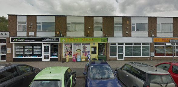 Row of shops with parked cars, featuring a local convenience store, under a cloudy sky