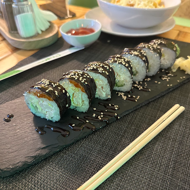 Sushi rolls with sesame seeds on slate, accompanied by chopsticks and a dish of sauce, served on a dining table