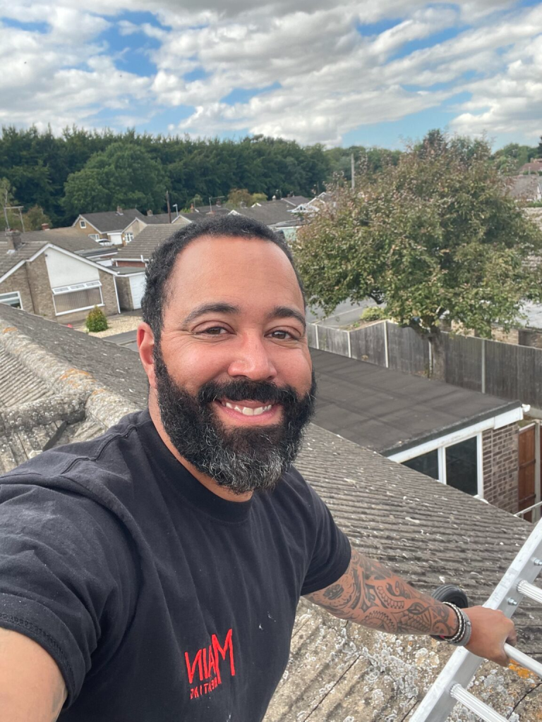Man with beard smiling on rooftop, holding ladder, surrounded by houses and trees under a cloudy sky