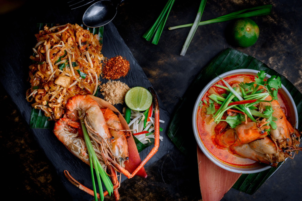 Thai cuisine with Pad Thai and Tom Yum soup garnished with prawns, lime, and fresh herbs on dark background