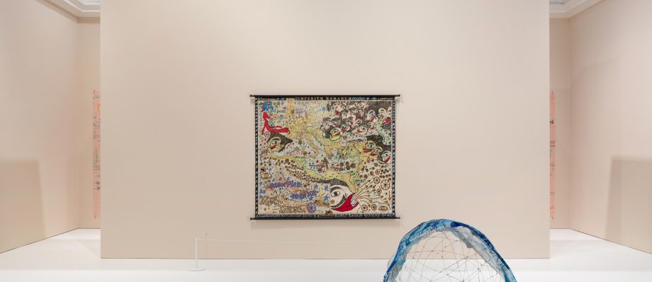 Contemporary art installation with a colourful map tapestry and a blue-accented sculpture in a minimalist gallery setting