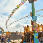 Colorful outdoor festival scene featuring a Ferris wheel, flags, and a vibrant crowd enjoying the sunny event.