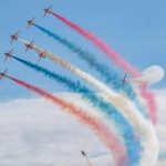 Seven planes perform aerobatics with vibrant smoke trails in the sky