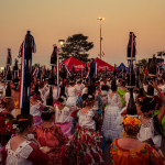 Traditional dance festival at sunset with women in colourful dresses and flower crowns, holding bottles in a lively celebration