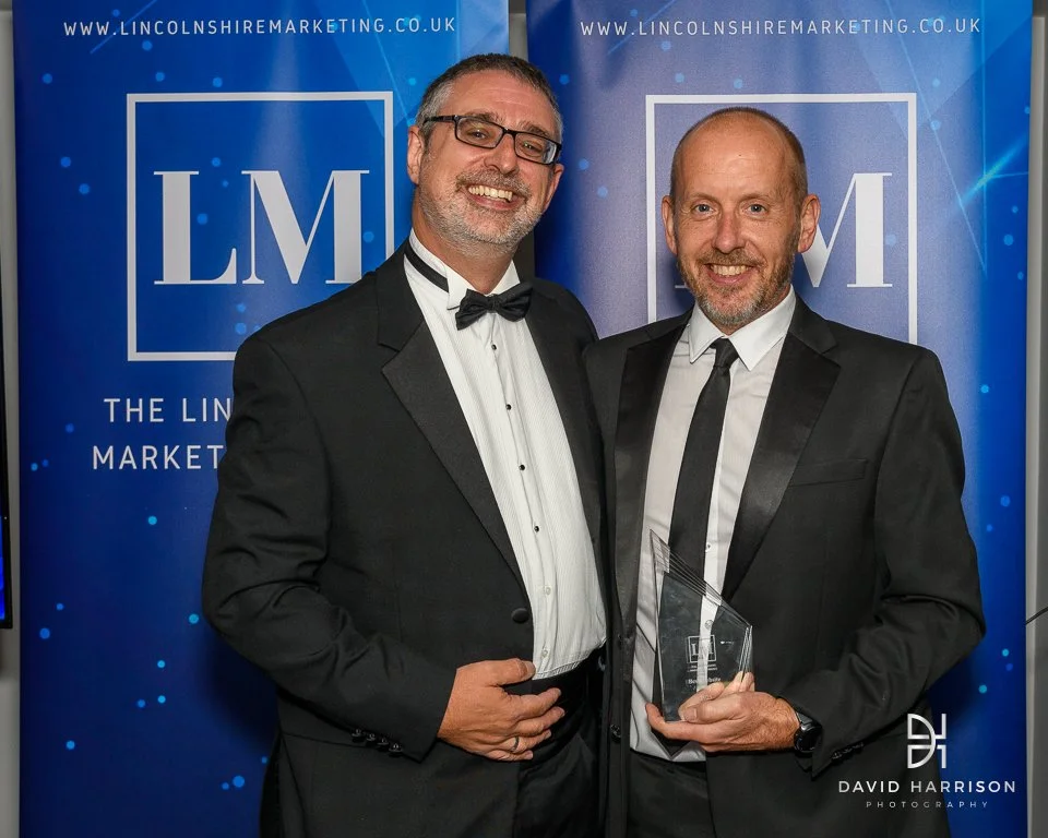 Two men in tuxedos at Lincolnshire Marketing event, one holding an award, with branded banners in the background
