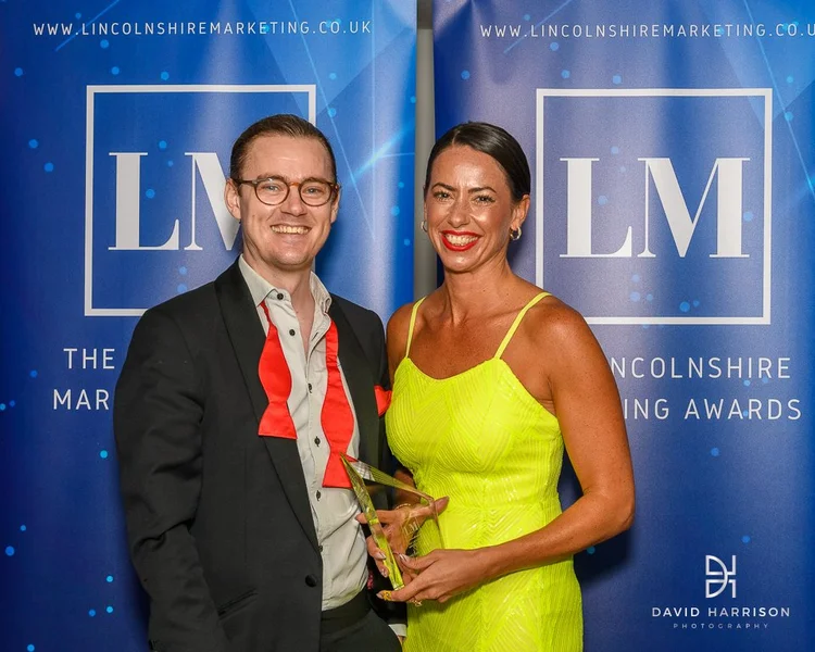 Two people smiling and holding an award at the Lincolnshire Marketing Awards event with blue banners in the background