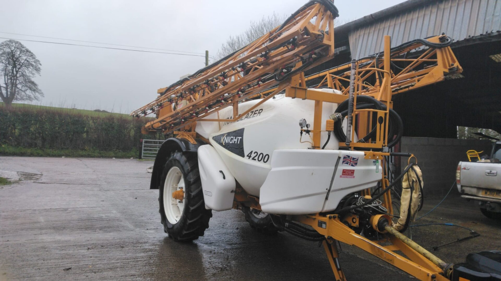 Agricultural sprayer with Knight 4200 model parked on a farm near a metal shed, surrounded by green fields