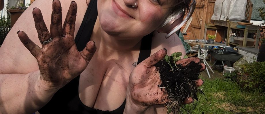 Person smiling while gardening, showing dirt-covered hands and small plants outside a brick house, wearing headphones