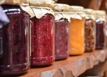 Row of assorted homemade jams in glass jars with paper lids on a wooden shelf