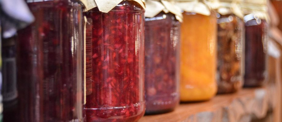 Row of assorted homemade jams in glass jars with paper lids on a wooden shelf