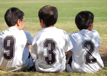 Three young boys in white football jerseys numbered 9, 3, and 2 seated on the grass field during daylight