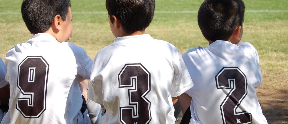 Three young boys in white football jerseys numbered 9, 3, and 2 seated on the grass field during daylight