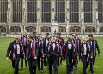 Group of young men in suits with striped scarves walk on grass in front of historic building with arched windows