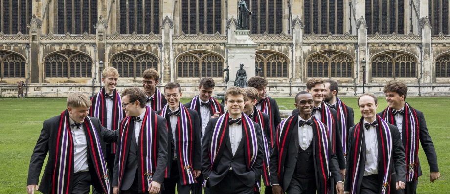 Group of young men in suits with striped scarves walk on grass in front of historic building with arched windows