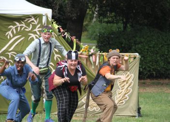 Four performers in playful costumes acting in an outdoor theatre production with green nature backdrop