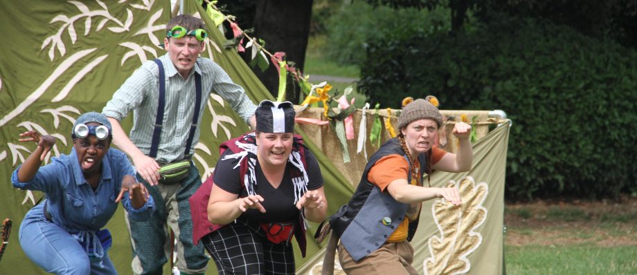 Four performers in playful costumes acting in an outdoor theatre production with green nature backdrop