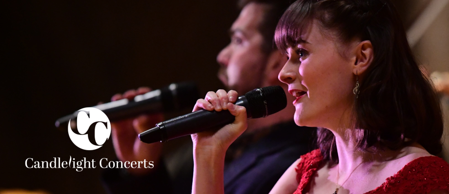 Female singer performing at Candlelight Concerts, holding microphone, in a red dress with male singer in the background