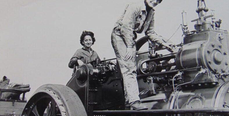 Two women operating a vintage steam traction engine in black and white, showcasing historical engineering skills