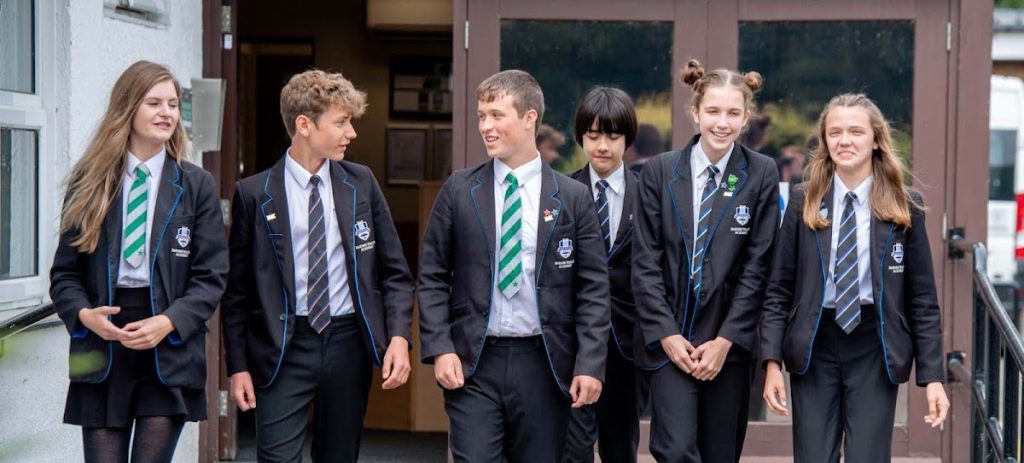 Group of six students in school uniforms walking outside a building, smiling and engaged in conversation