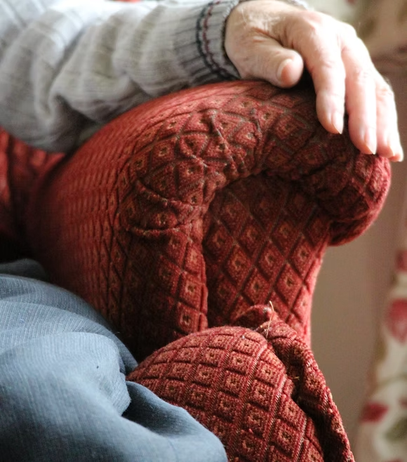 Elderly hand resting on a patterned red armchair, symbolising comfort and relaxation in a cosy home setting