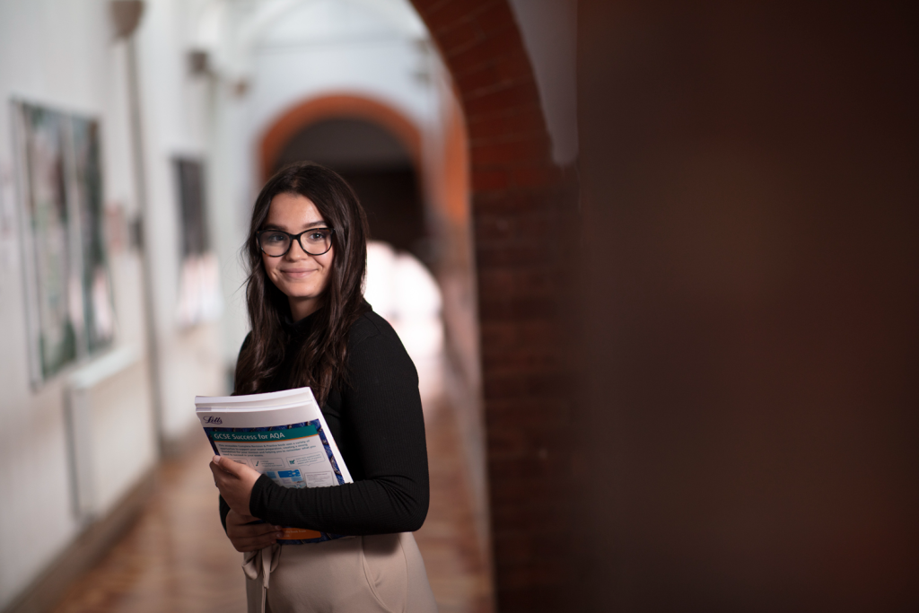 Student smiling in a school hallway holding study materials, wearing glasses and a black top, ready for exams