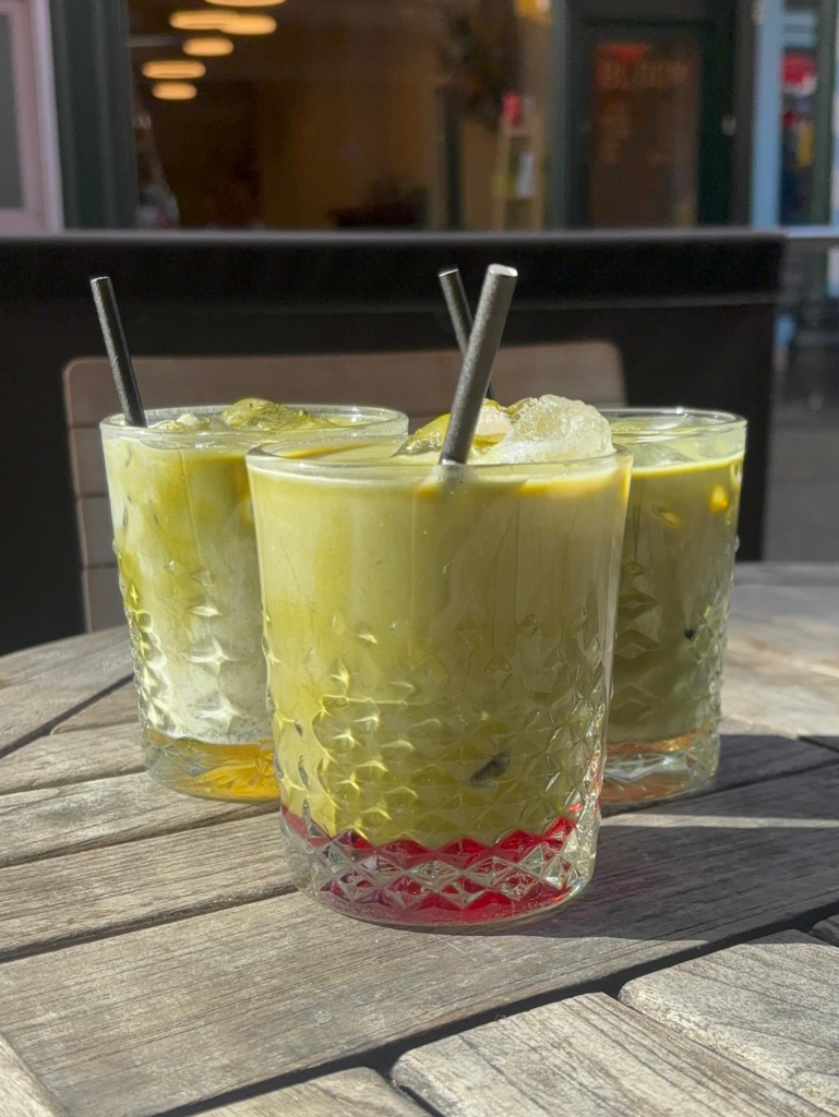Three colourful layered drinks in textured glasses with metal straws on a wooden table in an outdoor café setting