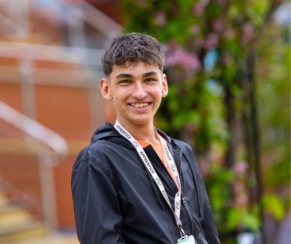 Smiling person standing outdoors, wearing a lanyard and black jacket, with greenery and blurred background