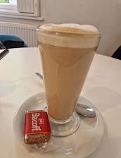 Latte in a tall glass with a foamy top, served with a Biscoff biscuit on a white table in a cosy cafe setting