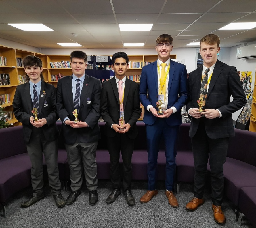 Five students in formal attire holding awards in a library setting, celebrating an achievement