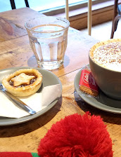 Cappuccino with biscuit, a mince pie on a plate, and a glass of water on a wooden café table near a cosy red scarf