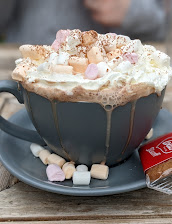 Grey mug of hot chocolate topped with whipped cream and marshmallows, served with a biscuit on a wooden table