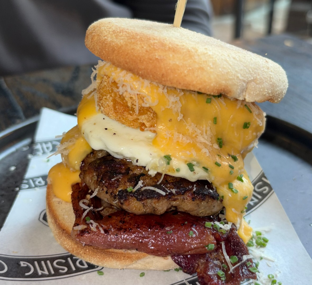 Stacked gourmet burger with cheese, bacon, and a crispy onion ring, served on a wooden table at a restaurant