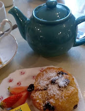 Tea time setup with green teapot, blueberry scone dusted with icing sugar, and fresh strawberries on a plate