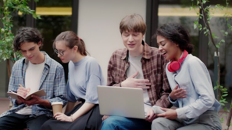 Group of diverse students enjoying a study session outdoors, with a laptop and notebook, surrounded by greenery