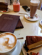 Café table with slices of cake, cappuccino, hot chocolate, and a mason jar drink, next to a closed menu on a wooden surface